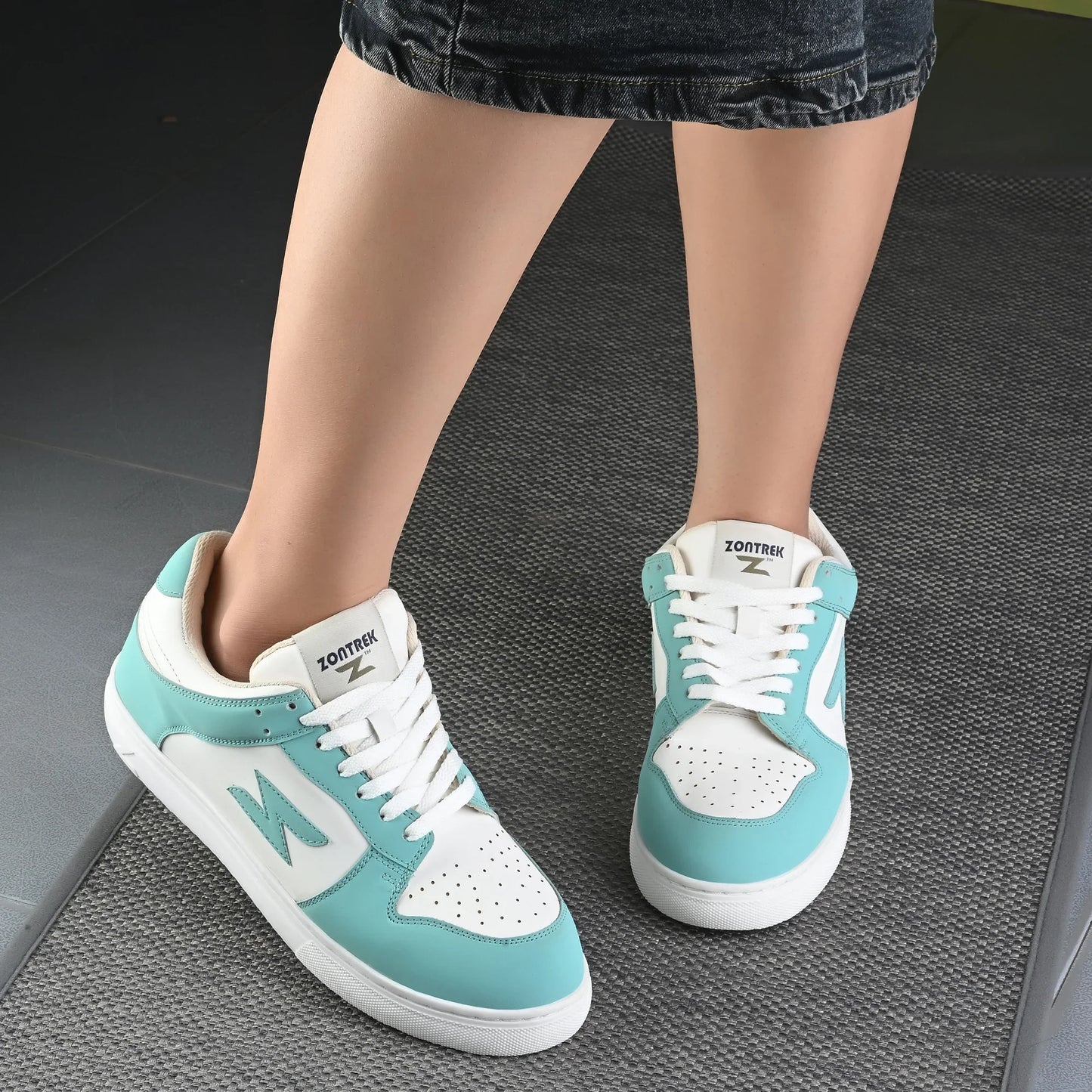 Teal and white sneakers worn by a person on a dark textured floor.